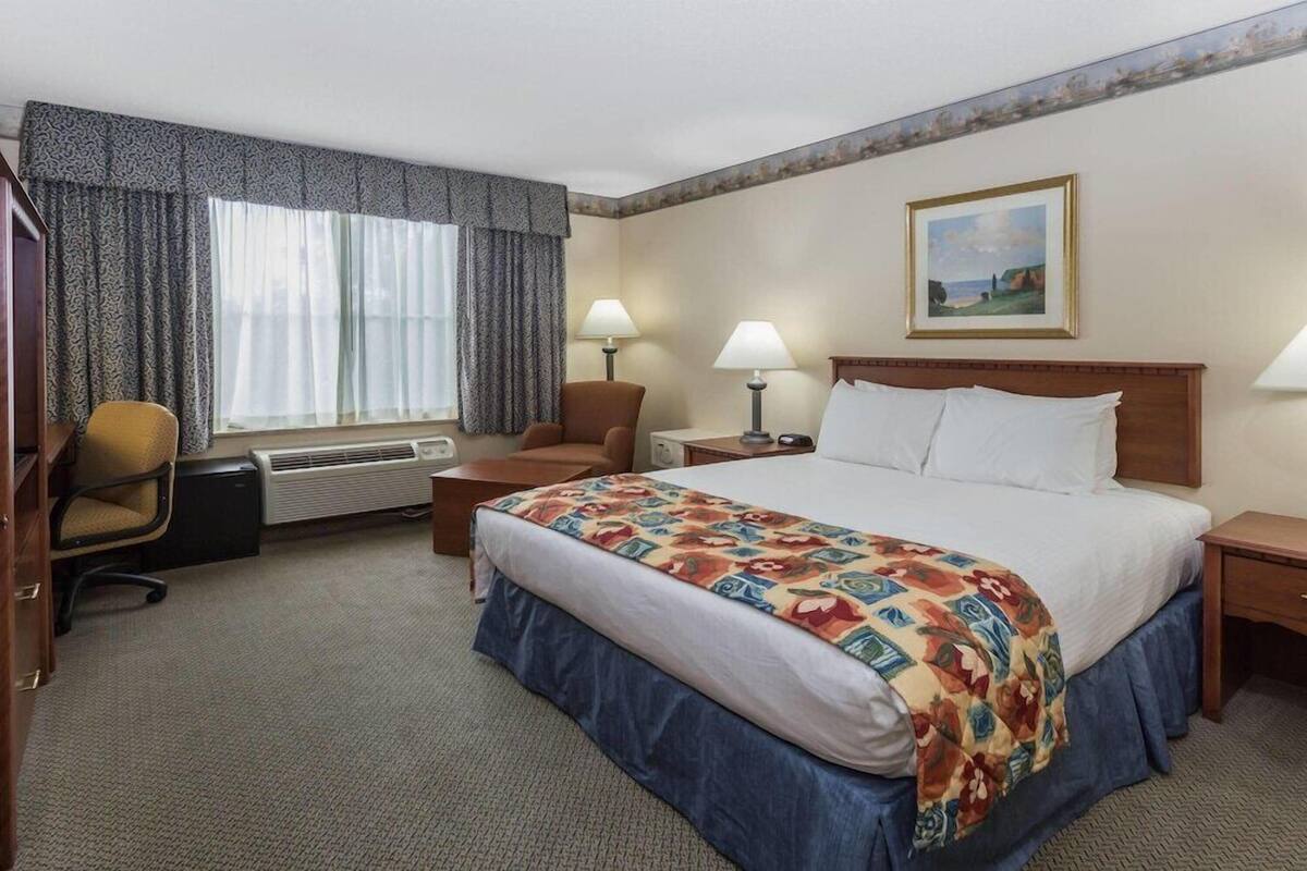 Habitación de hotel con cama, dos lámparas de noche, una silla, un escritorio y un ventanal con cortinas.