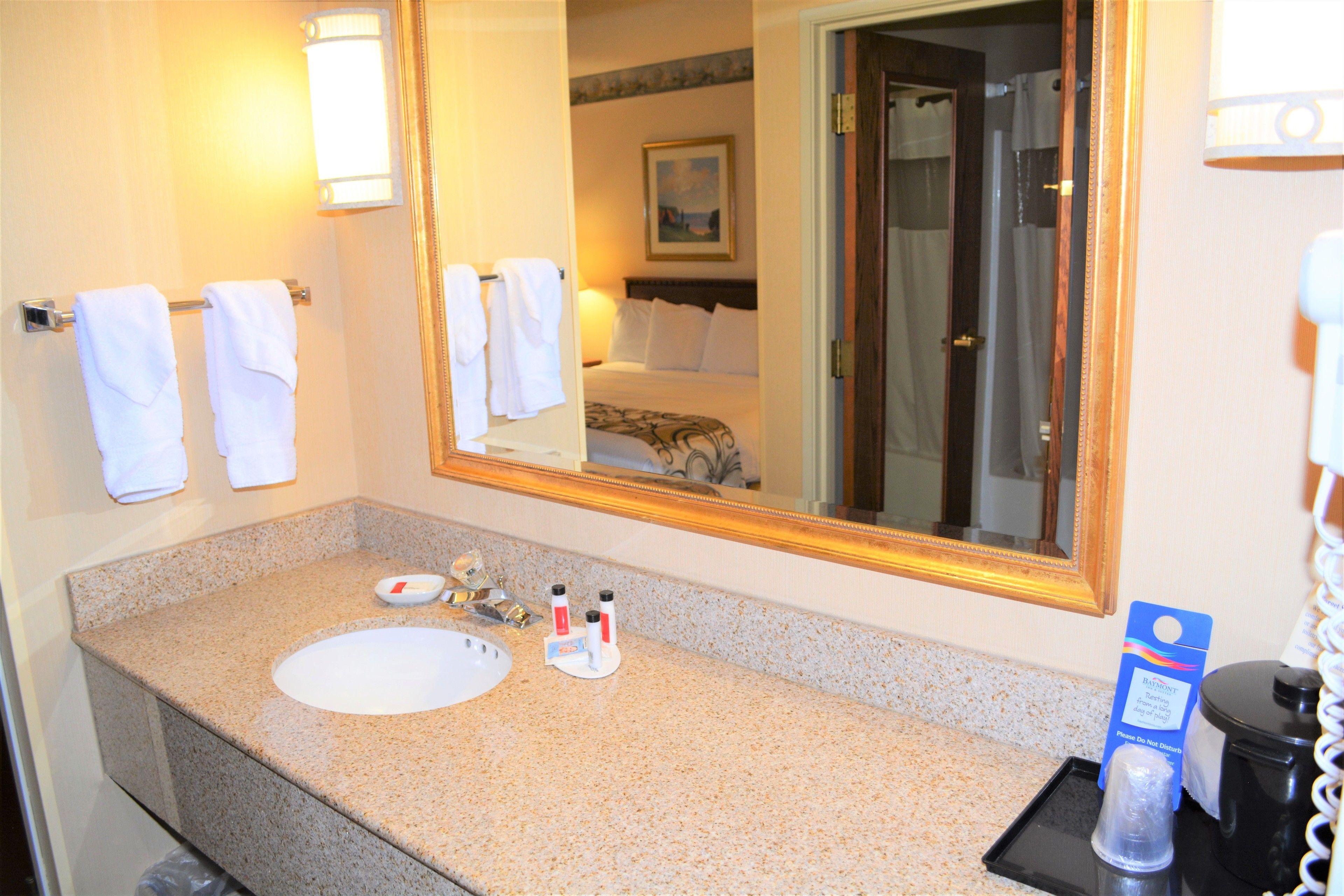 A hotel bathroom with a granite countertop sink, a large mirror, and a bed visible in the reflection.