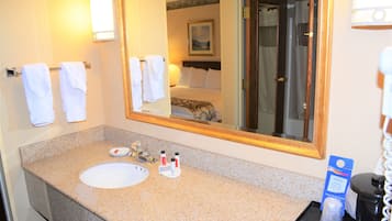 A hotel bathroom with a granite countertop sink, a large mirror, and a bed visible in the reflection.