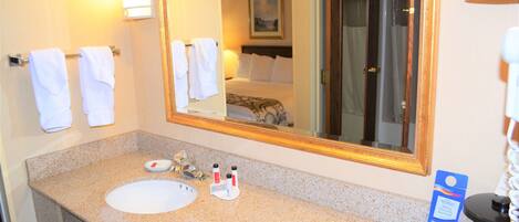 A hotel bathroom with a granite countertop sink, a large mirror, and a bed visible in the reflection.