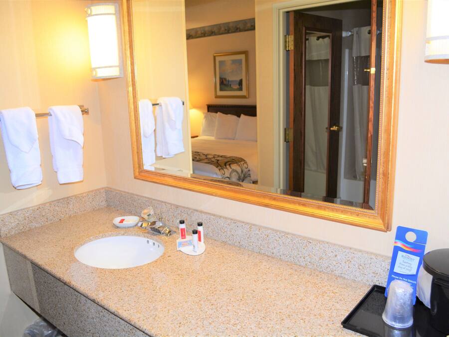 A hotel bathroom with a granite countertop sink, a large mirror, and a bed visible in the reflection.