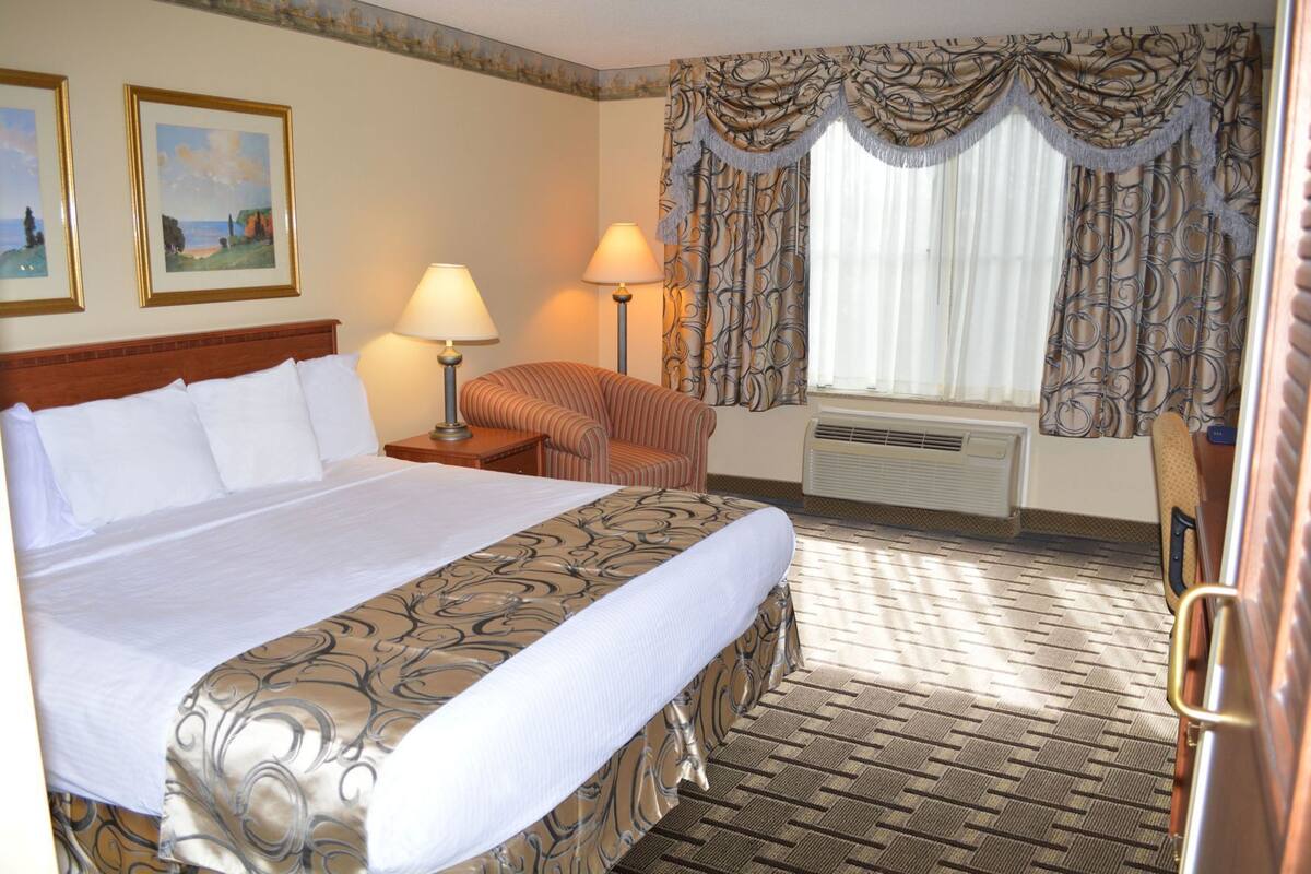 Habitación de hotel con una cama grande, dos lámparas de noche, un sillón a rayas, una ventana con cortinas y un radiador.
