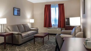 Quarto de hotel com sofá bege, uma pequena mesa, televisão e janela com cortinas vermelhas.