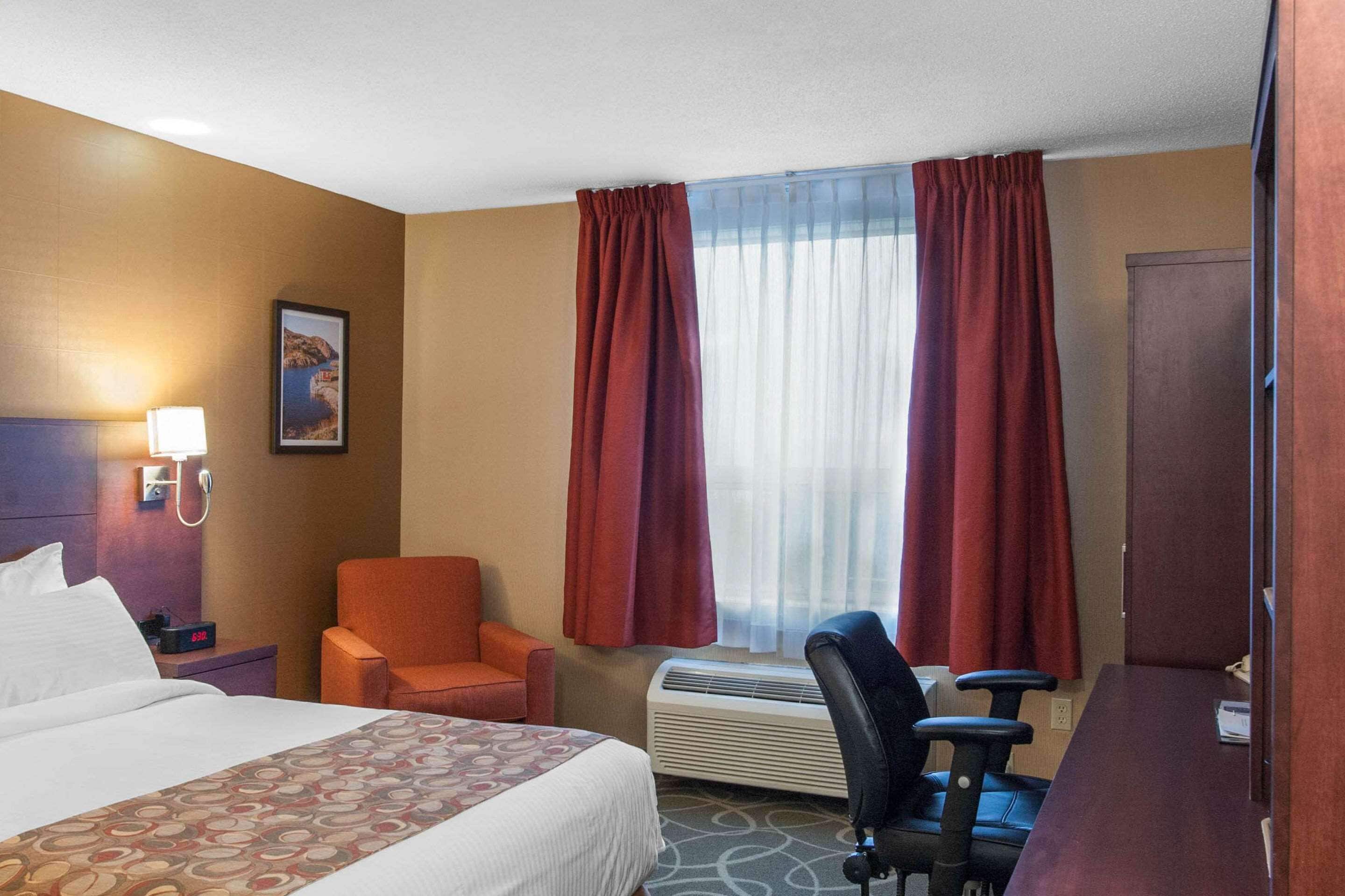 A hotel room with a bed, an orange chair, a desk with a chair, a wall-mounted lamp, a picture, and a window with red curtains.