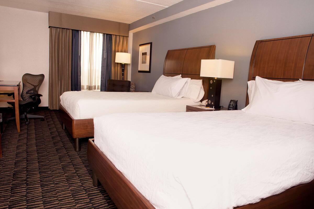 A hotel room with two beds, a desk, a chair, and a lamp.