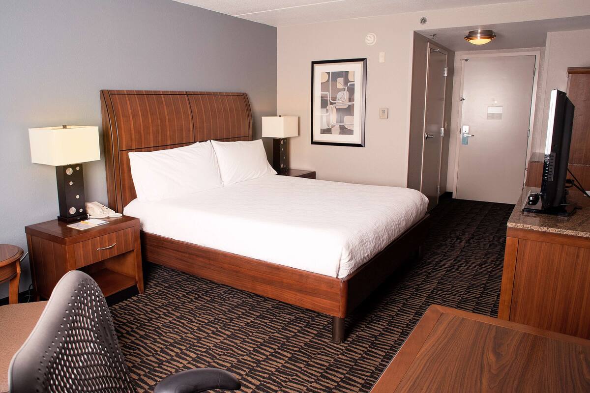 A hotel room with a wooden bed, two bedside lamps, a desk with a computer, and a television on a wooden stand.