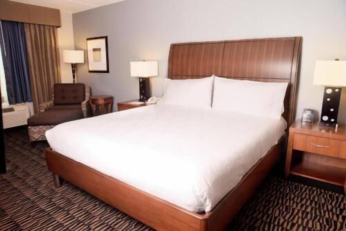 A hotel room with a large bed, a wooden headboard, a desk with a lamp, a chair, and a small table.