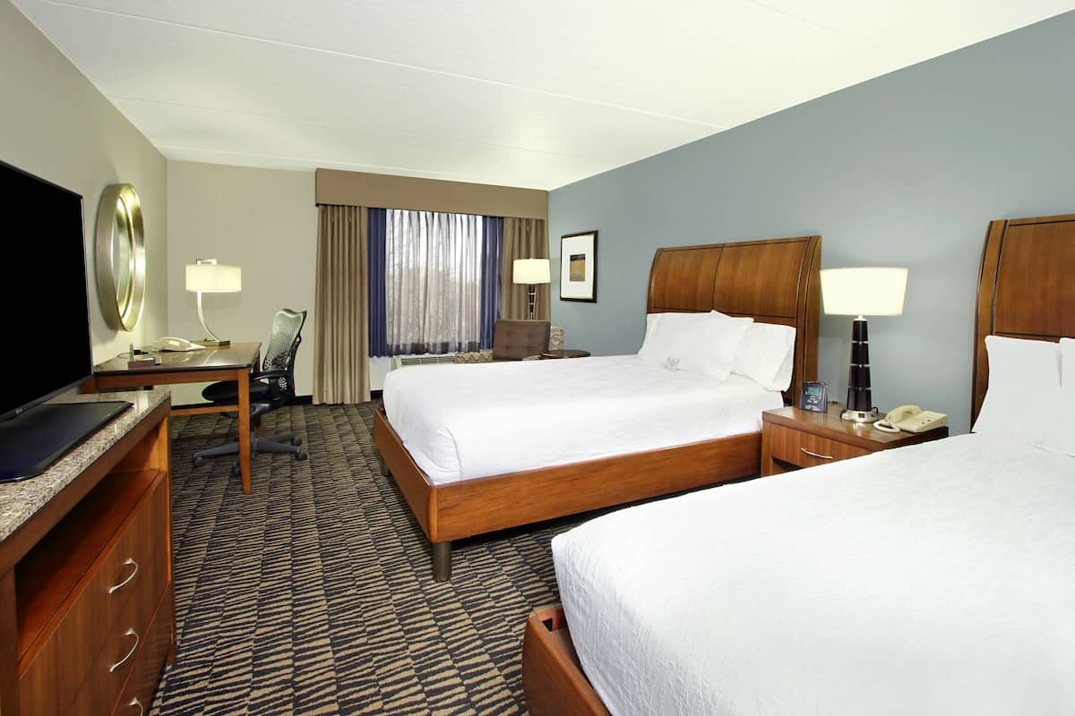 A hotel room with two beds, a desk, a chair, a television, and a window with curtains.