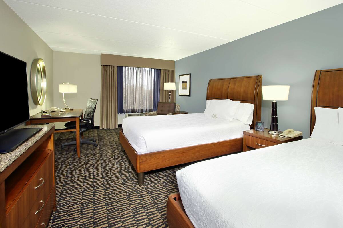 A hotel room with two beds, a desk, a chair, a TV, and a window with curtains.