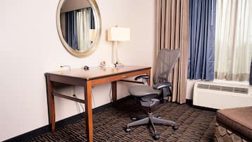 Habitación de hotel con un escritorio de madera, una silla de oficina negra, un espejo redondo, una lámpara, un teléfono, una alfombra estampada, una ventana con cortinas y un sofá.