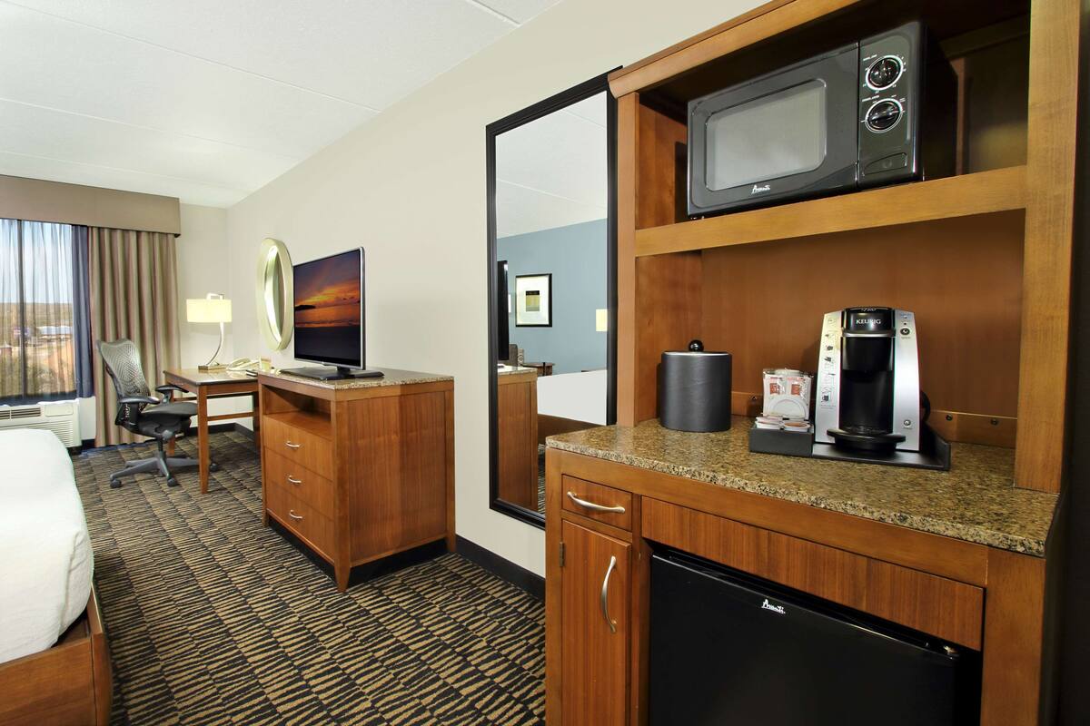 A hotel room with a bed, desk, chair, TV, microwave, coffee maker, and a small refrigerator.