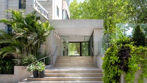 Entrada moderna de um edifício com escadaria que leva a uma porta de vidro, cercada por vegetação e plantas em vasos.