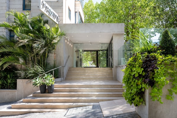 Una entrada moderna a un edificio con una escalera que conduce a una puerta de cristal, rodeada de vegetación y plantas en macetas.