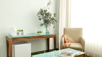 A compact room with a wooden desk, a small refrigerator, a glass coffee table, a beige armchair, and a potted plant.