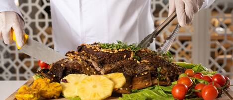 A chef slicing a large, seasoned meat dish on a wooden board, garnished with pineapples, tomatoes, and lettuce.