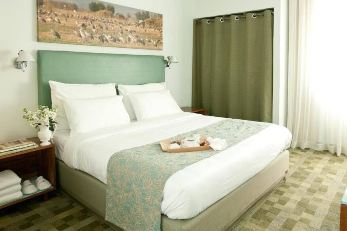 Una cama bien hecha con un cabecero verde, una mesita de noche con una lámpara y una bandeja con tetera y tazas.