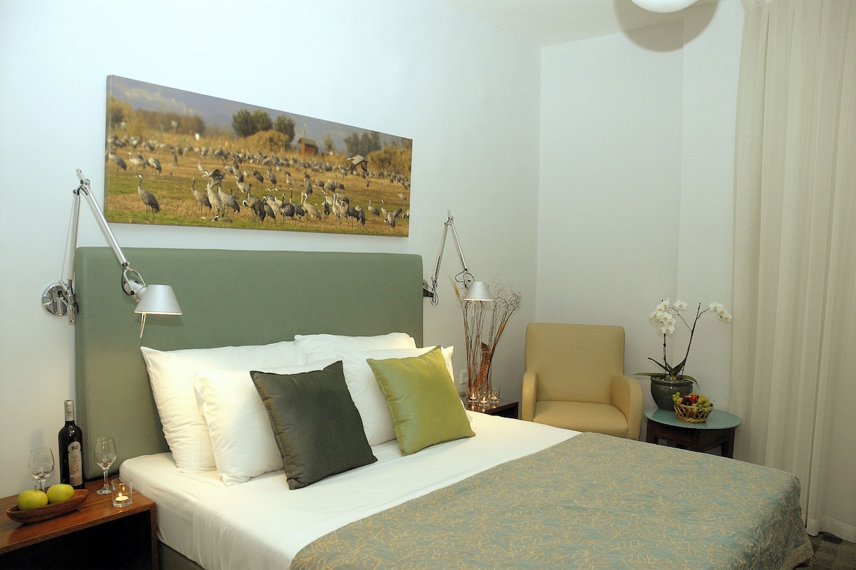 Una cama bien hecha con almohadas, una mesita de noche con una botella y vasos, una silla, un jarrón con flores y un cuadro de pájaros en la pared.