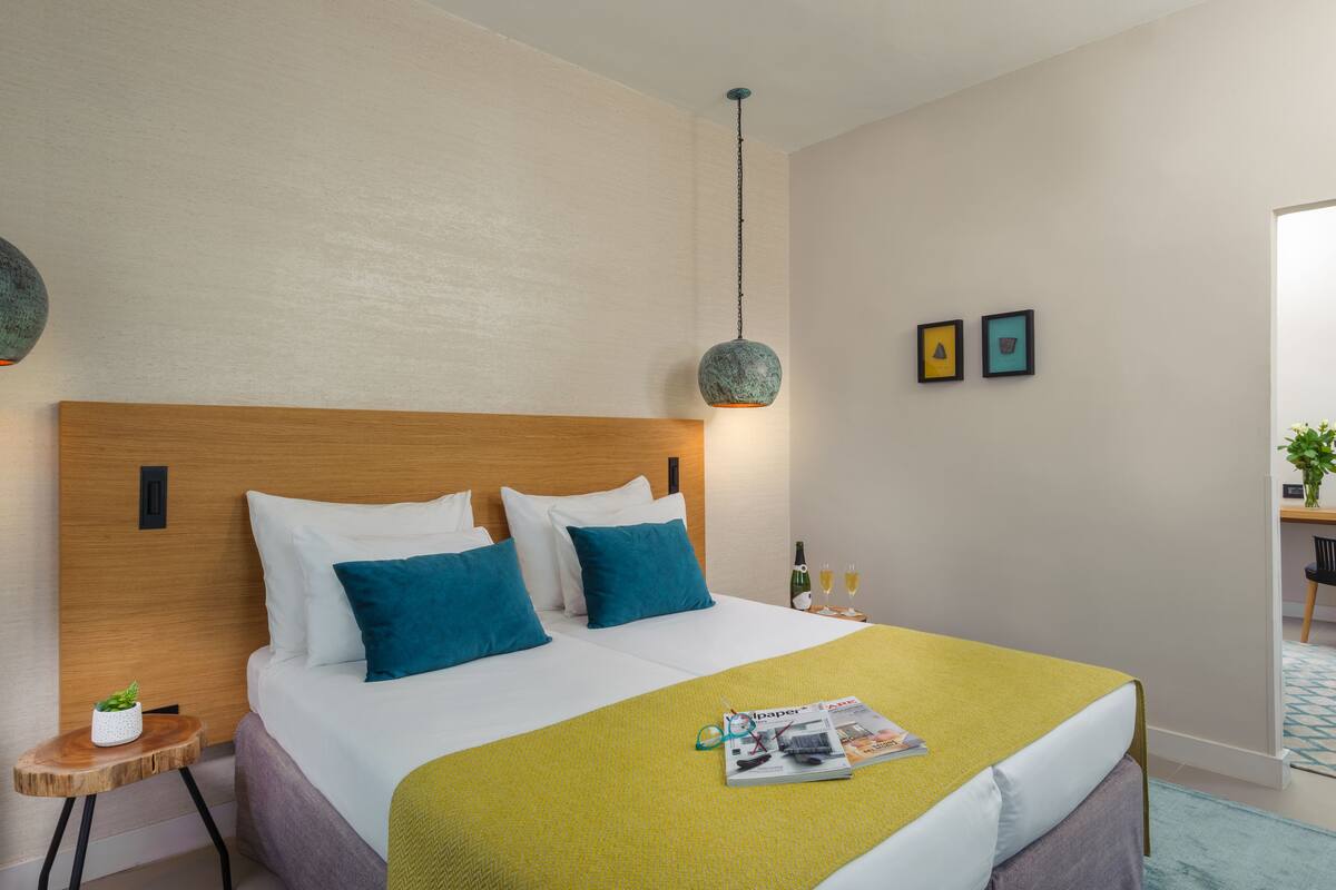 Habitaci ón de hotel con cabecera de madera, cama con almohadas blancas y verde azuladas, manta verde y una revista sobre la cama. Hay dos cuadros enmarcados en la pared y una mesita con una botella y vasos.