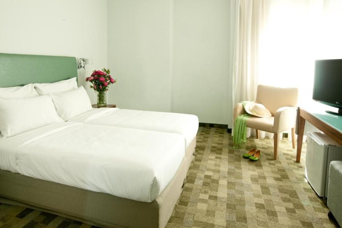 Habitación de hotel con cama, escritorio con televisor, una silla y una mesita con un jarrón de flores.