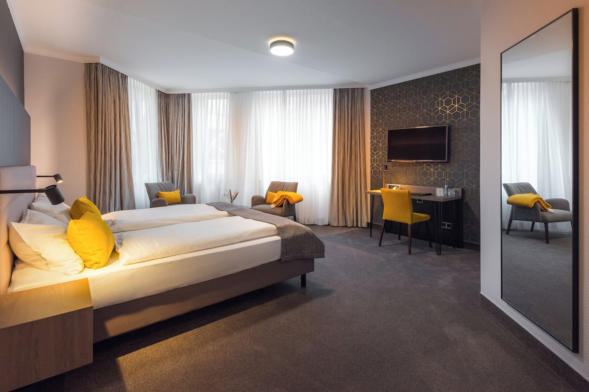 Quarto de hotel moderno com uma cama grande, uma escrivaninha, duas cadeiras, uma televisão e um espelho amplo.