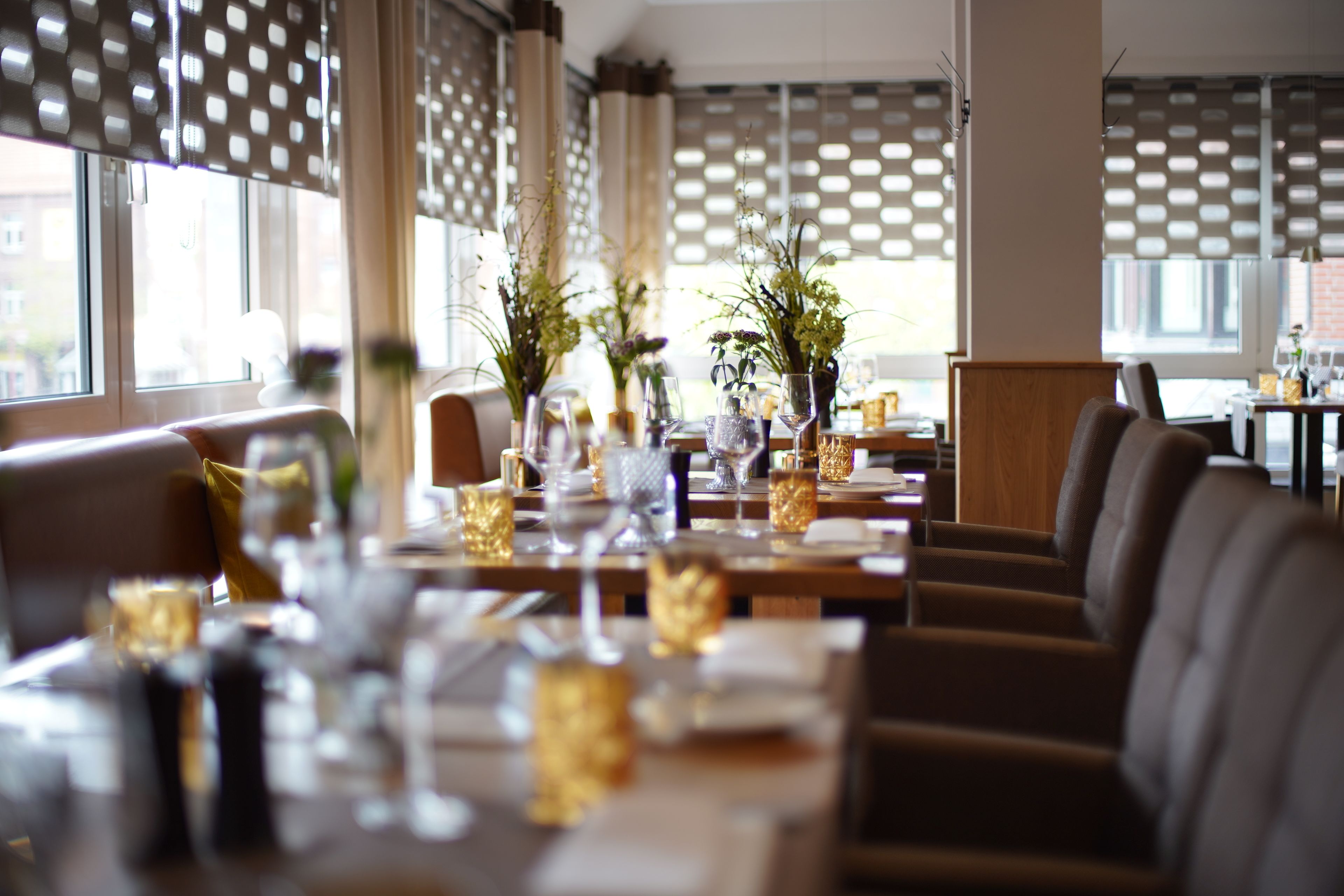 Restaurante com mesas preparadas para refeições, decoração moderna e luz natural de grandes janelas.