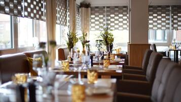 Restaurante com mesas preparadas para refeições, decoração moderna e luz natural de grandes janelas.