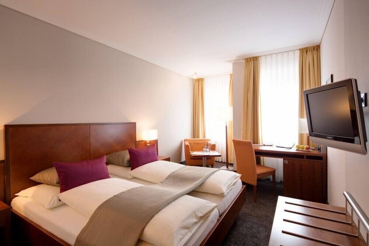 Quarto de hotel com cama, escrivaninha, cadeira, televisão e janela com cortinas.