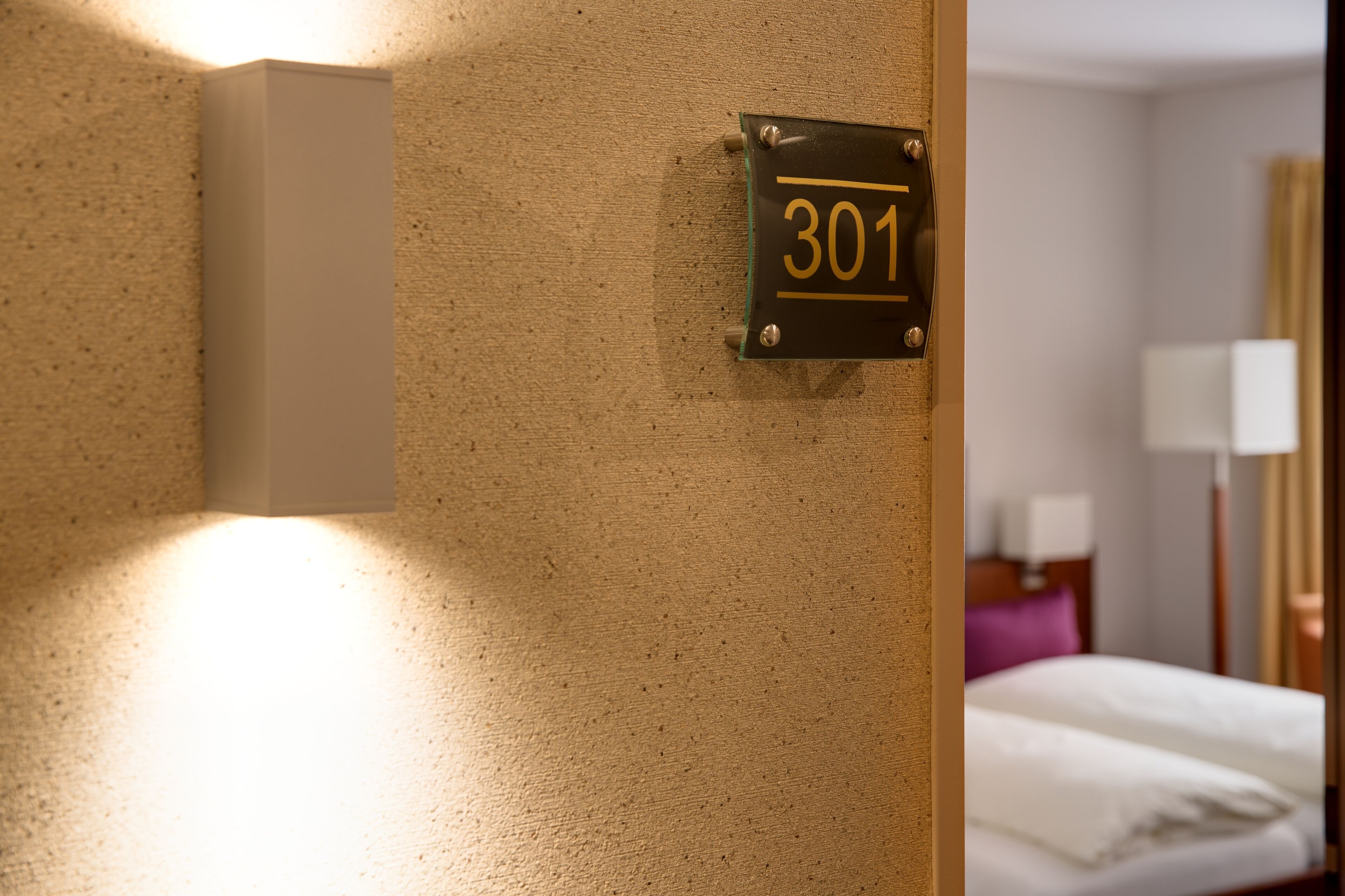 Una habitación de hotel con un cartel de número en la pared, una cama y una mesita de noche con una lámpara.