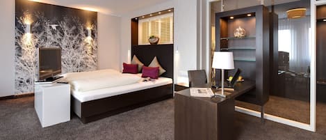 A modern hotel room with a bed, bedside table, and a desk with a lamp.
