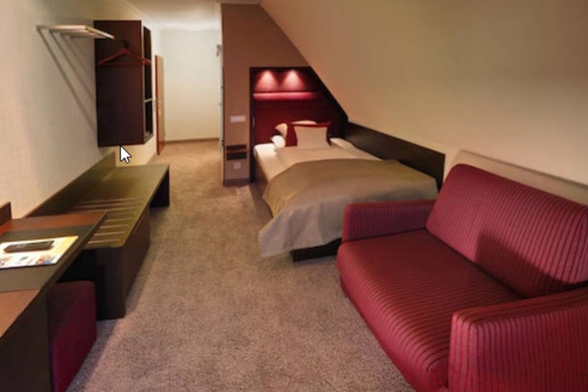Quarto de hotel pequeno, com uma cama, um sofá, uma escrivaninha e uma televisão.