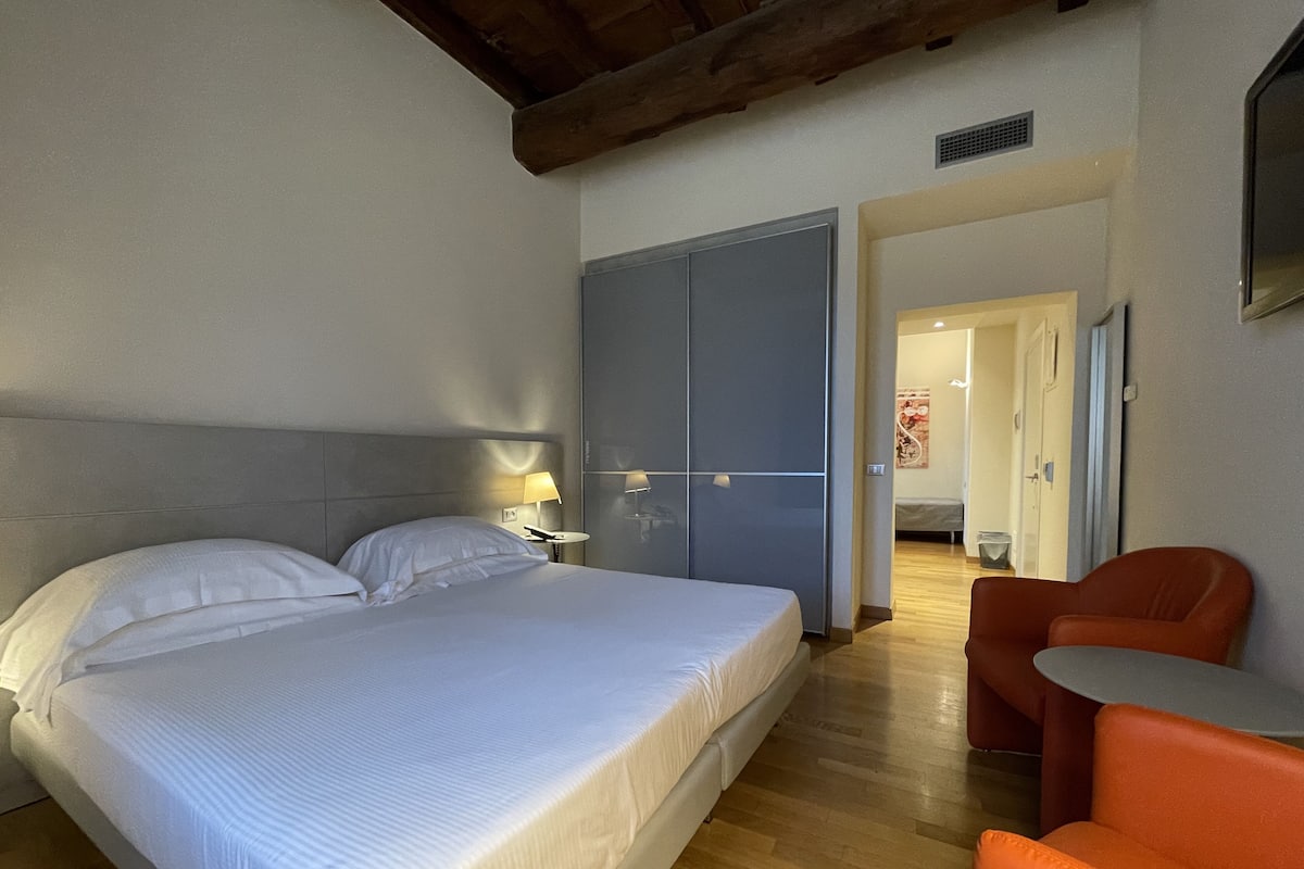 A hotel room with a bed, bedside lamps, a wardrobe, a small table, and a chair.
