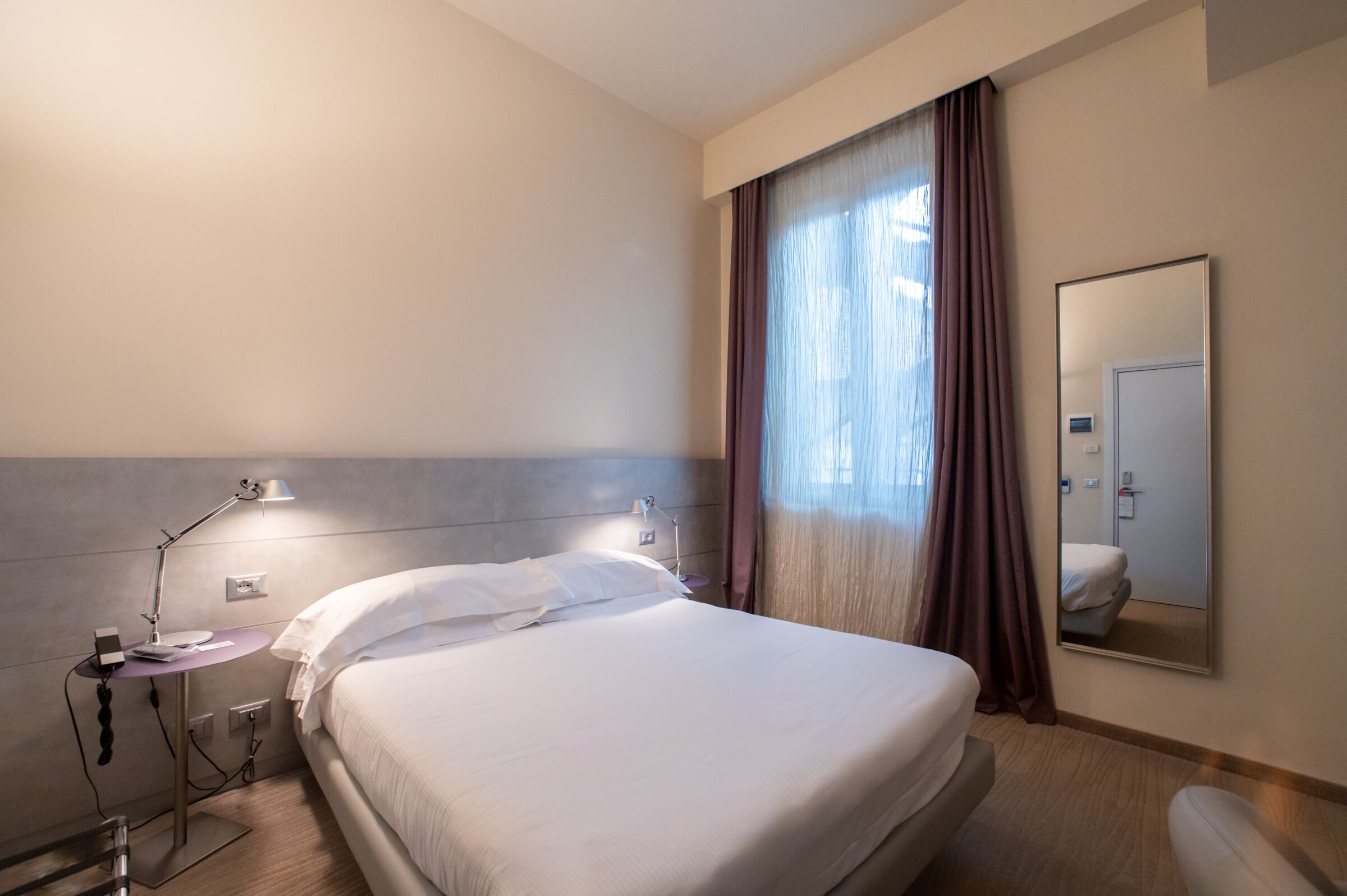 A hotel room with a bed, a bedside table with a lamp, a mirror, and a window with curtains.