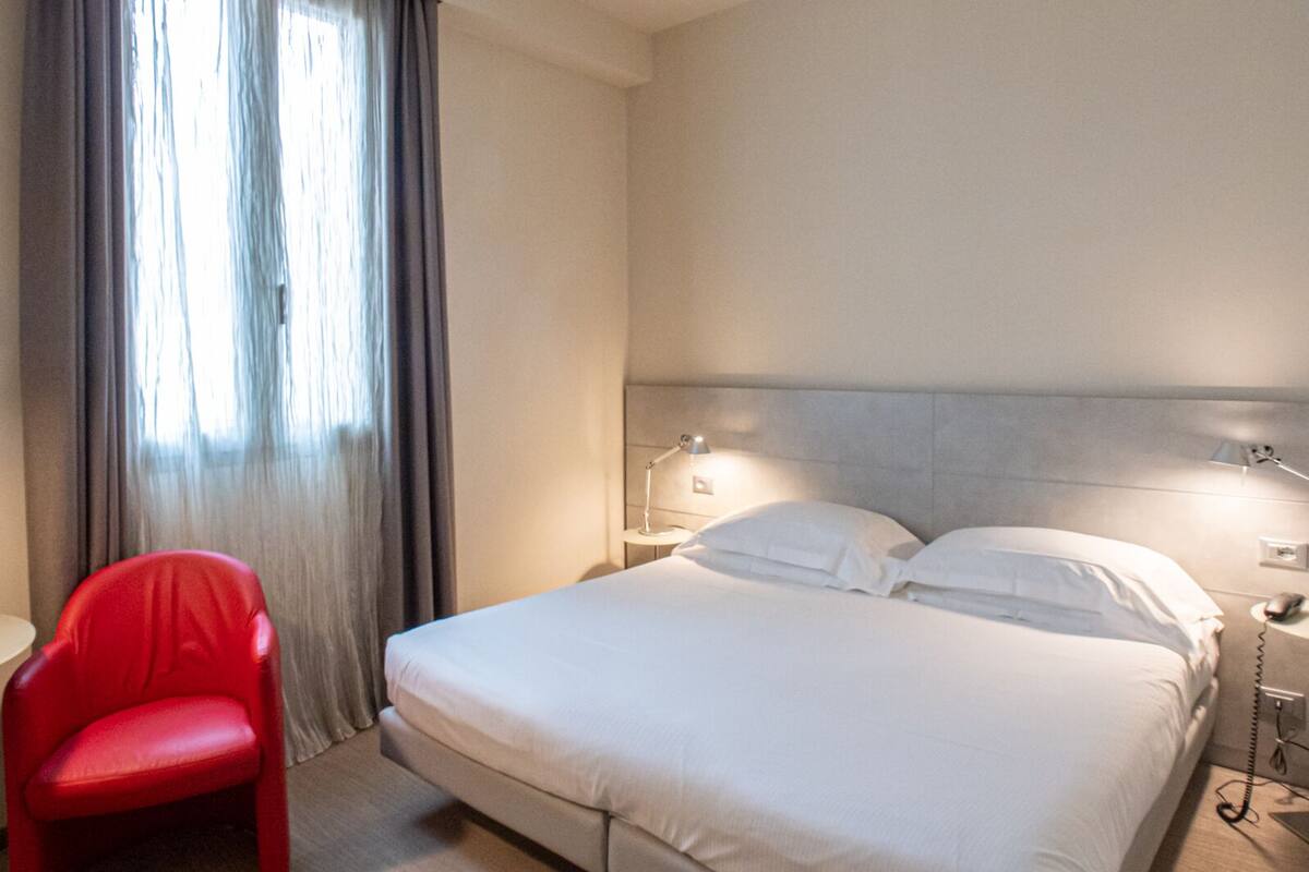 A hotel room with a bed, a red chair, a nightstand, and a window with curtains.