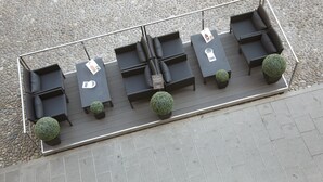 Una terraza en la azotea con una amplia zona de estar rectangular, con sofás negros y mesas, rodeada de pequeñas macetas.