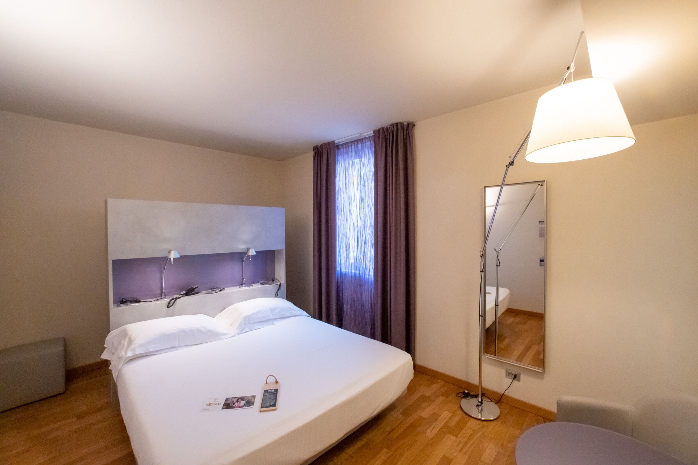 Ein Hotelzimmer mit einem großen Bett, einem Nachttisch mit Lampe, einem Spiegel und einem Fenster mit Vorhängen.