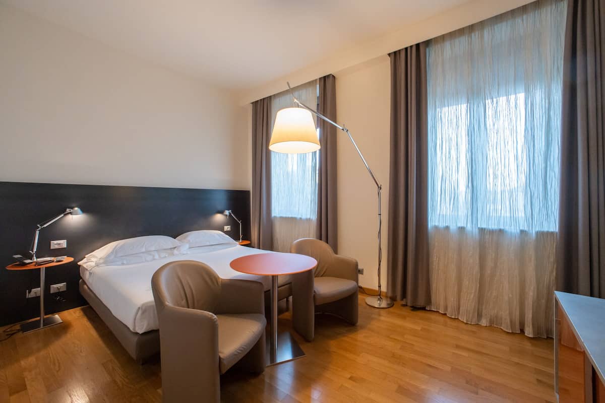 A hotel room with a bed, two bedside lamps, a round table, two chairs, and a large window with curtains.