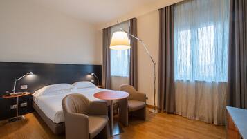A hotel room with a bed, two bedside lamps, a round table, two chairs, and a large window with curtains.