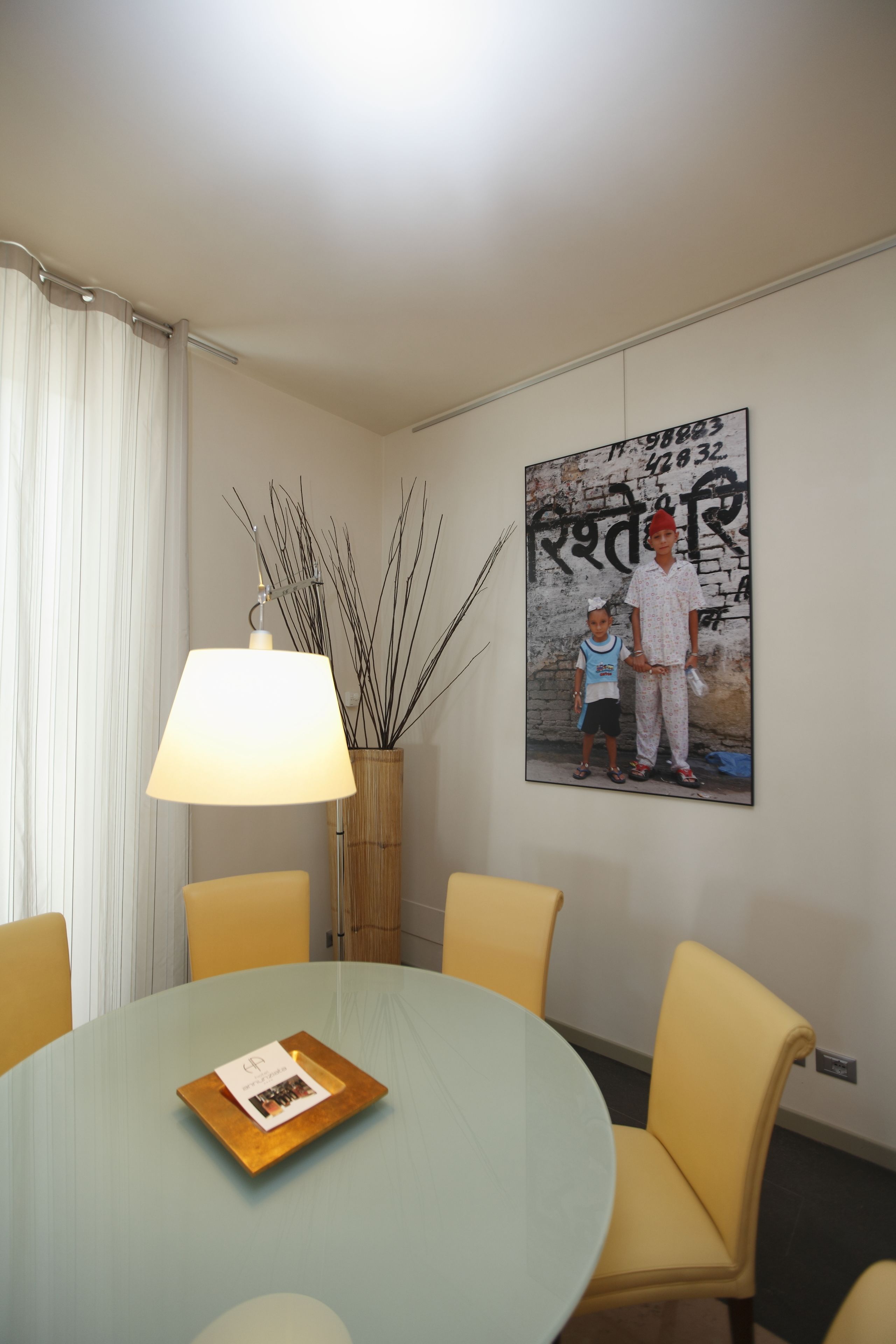 A dining area with a round table, yellow chairs, a lamp, a framed picture on the wall, and a small table with a menu.