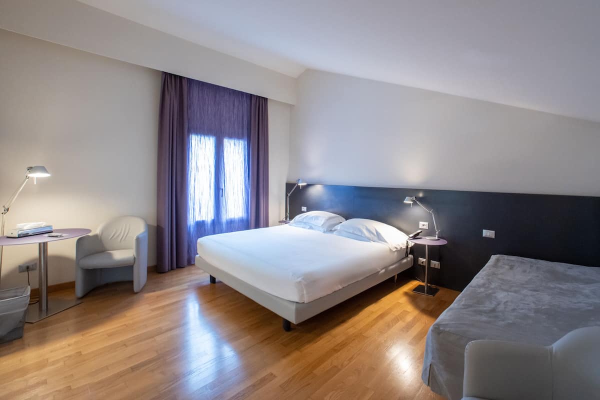 A hotel room with a bed, a desk, a chair, and a bedside table with lamps.