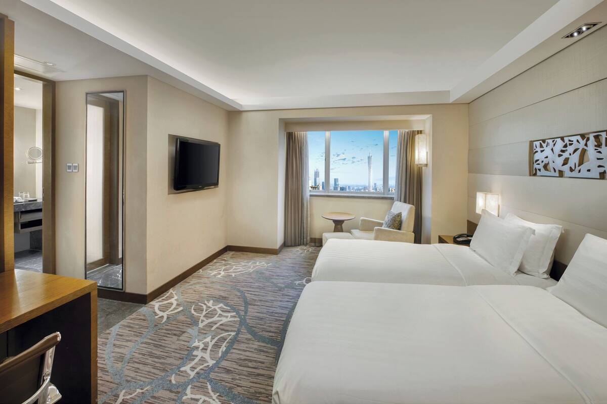A hotel room with two beds, a TV, a view of the cityscape, a small table, and a mirror.