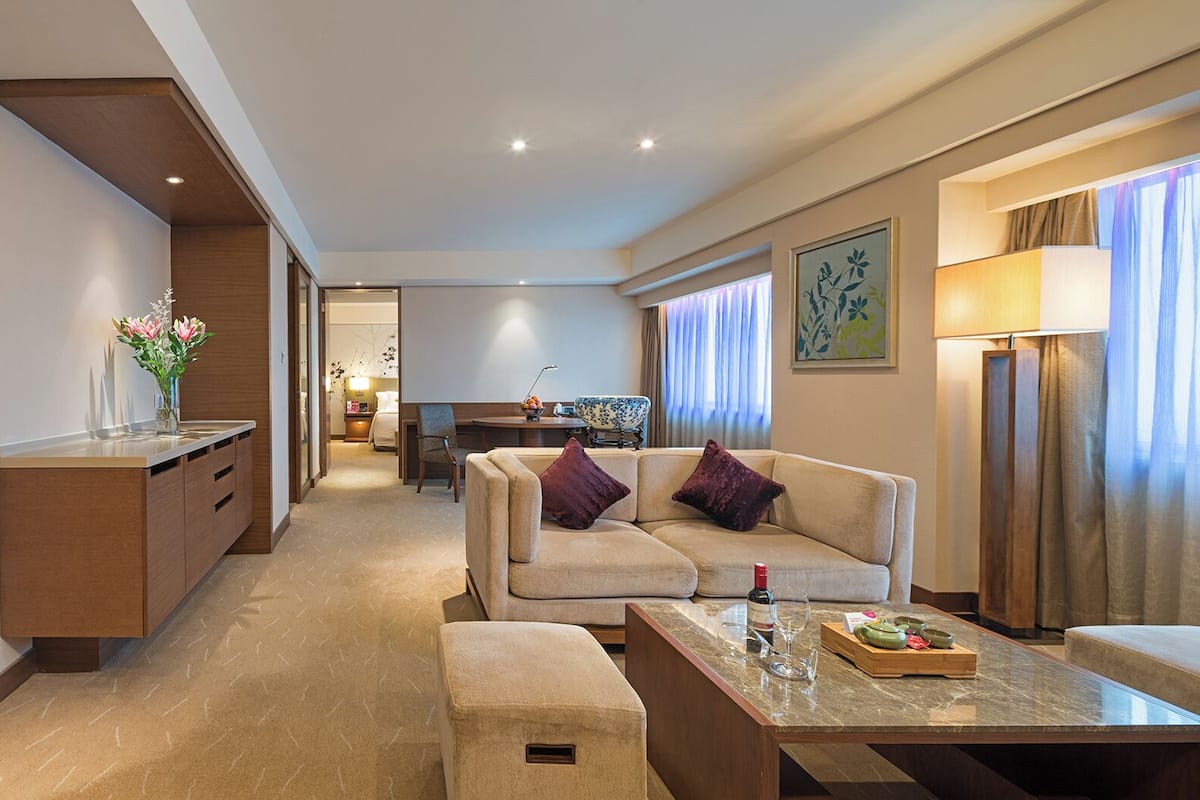 A modern hotel room with a sofa, armchair, and a dining table. There is a kitchen area in the background.