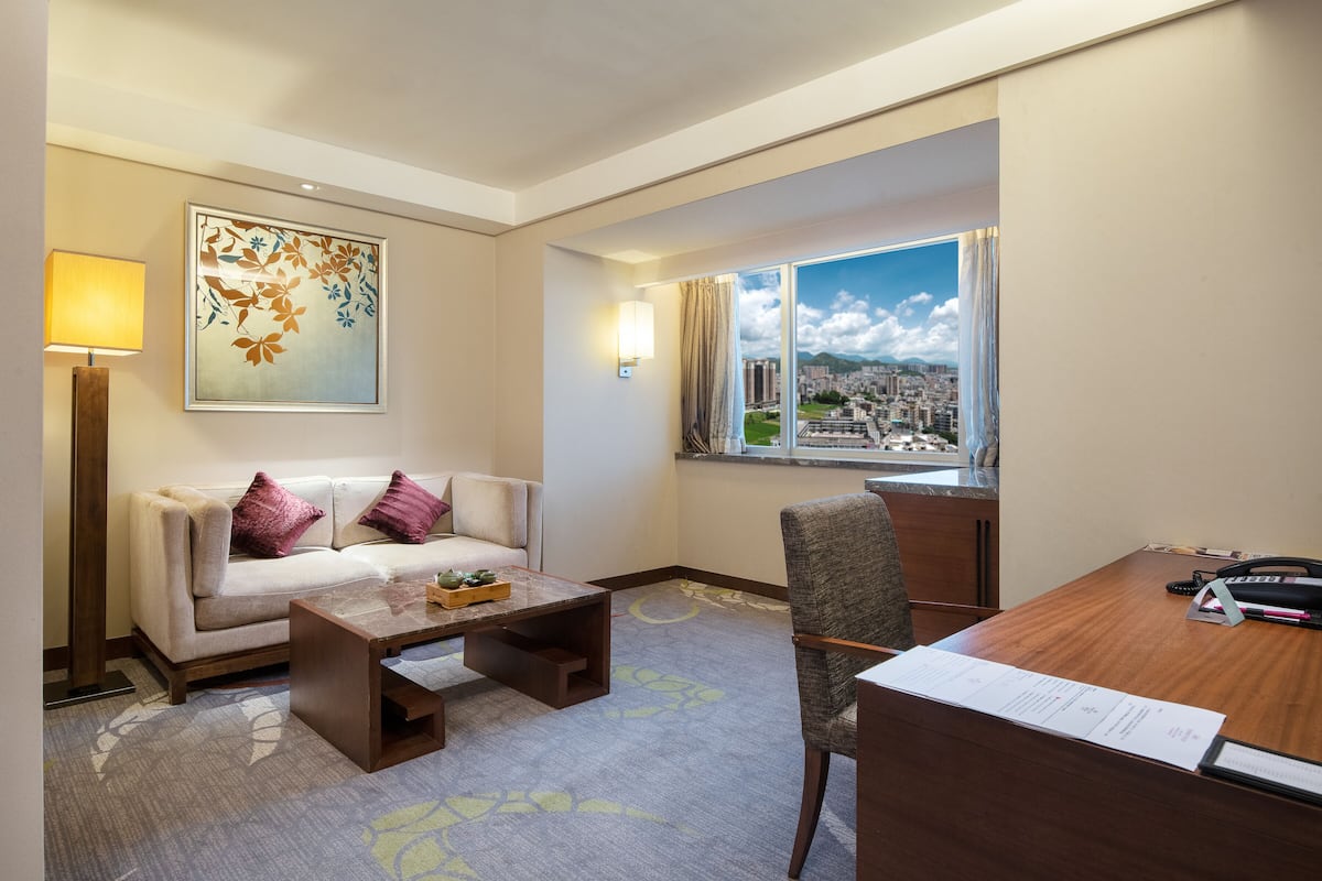 A hotel room with a sofa, a coffee table, a desk with papers, a lamp, a painting, and a window with a city view.