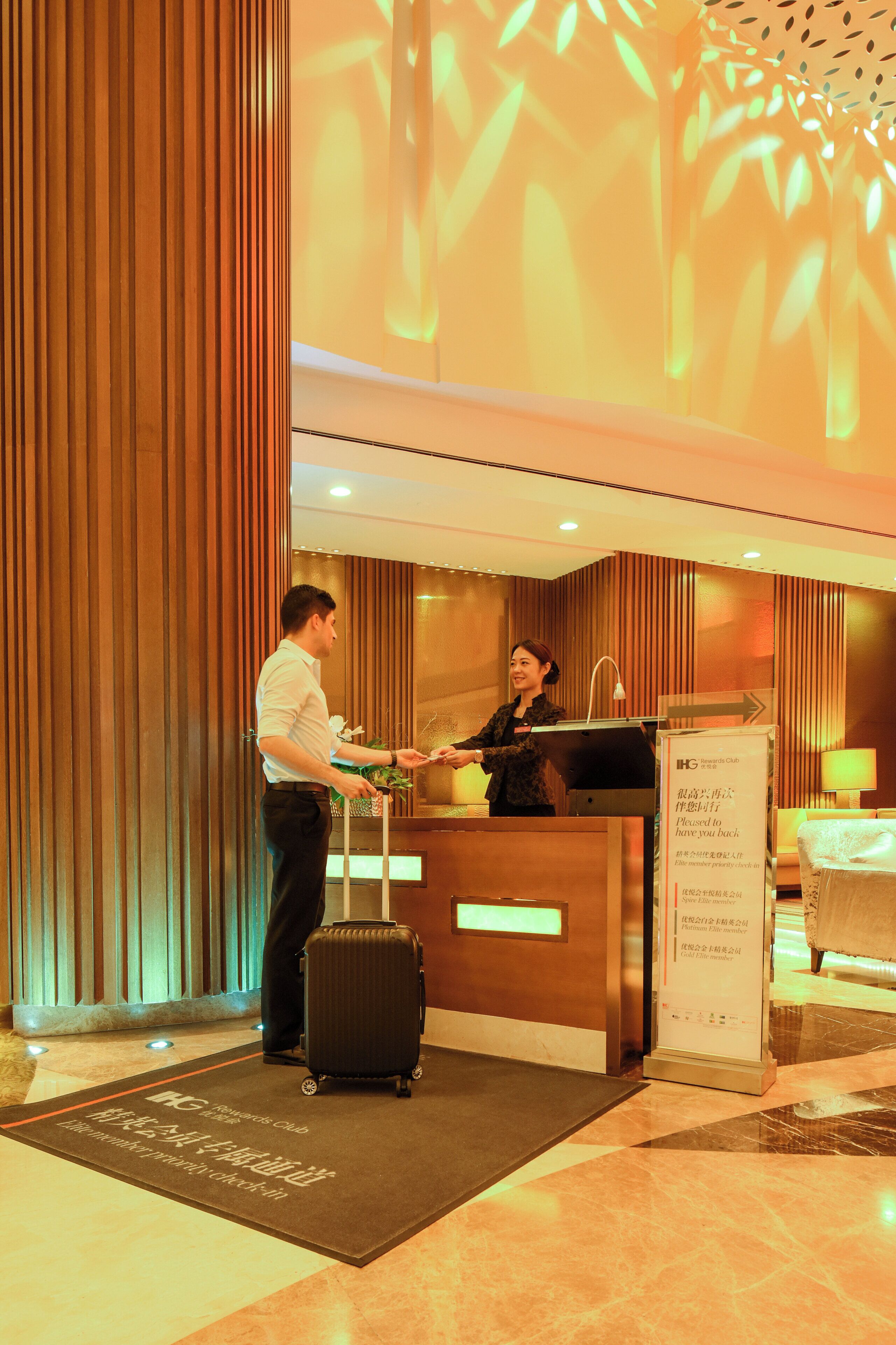 A hotel reception area with a reception desk, a guest with luggage, and a sign indicating the hotel is part of the IHG Rewards Club.