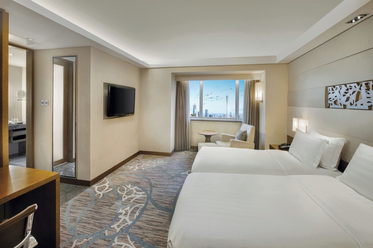 Habitación de hotel con dos camas, televisión, vista a la ciudad, una mesita y un espejo.