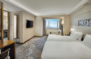 A hotel room with two beds, a TV, a view of the cityscape, a small table, and a mirror.