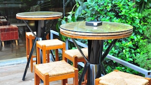 A set of round tables with woven chairs, positioned near a glass wall with a view of indoor greenery.