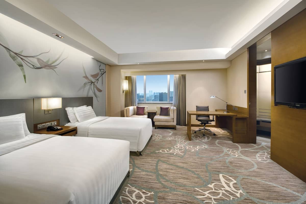 A hotel room with two beds, a desk, a chair, a TV, and a large window with a city view.
