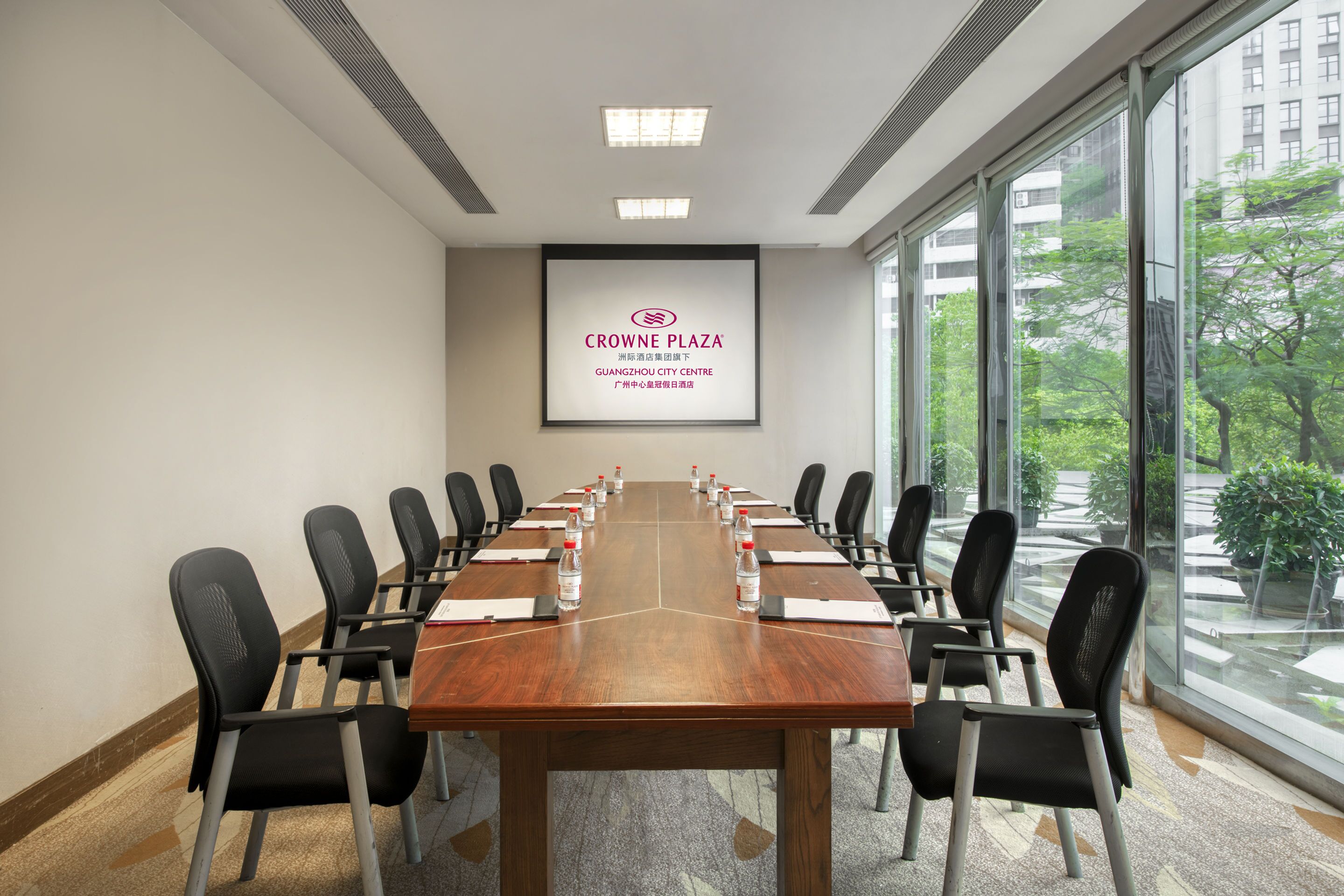 A conference room with a long table, chairs, a projector screen, and a view of greenery outside.