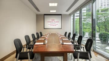 A conference room with a long table, chairs, a projector screen, and a view of greenery outside.