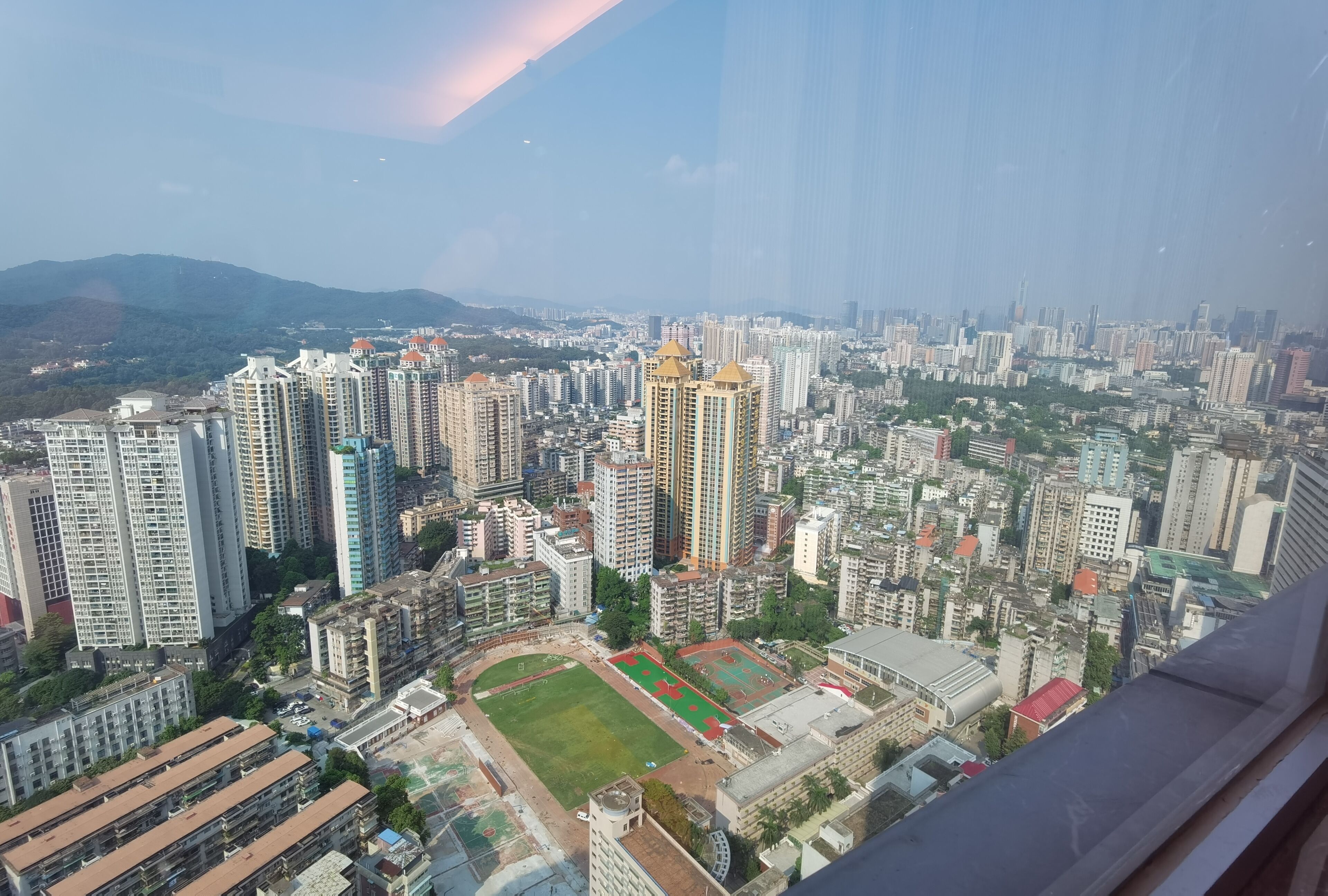 A high-rise building with multiple floors, surrounded by other high-rise buildings and a green area with sports fields.